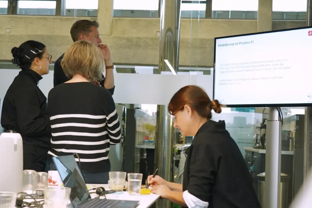 Das Team analysiert den IST-Prozess anhand einer Beispielanfrage. Auf dem Bildschirm ist eine typische Kundenanfrage für eine Konferenz zu sehen.